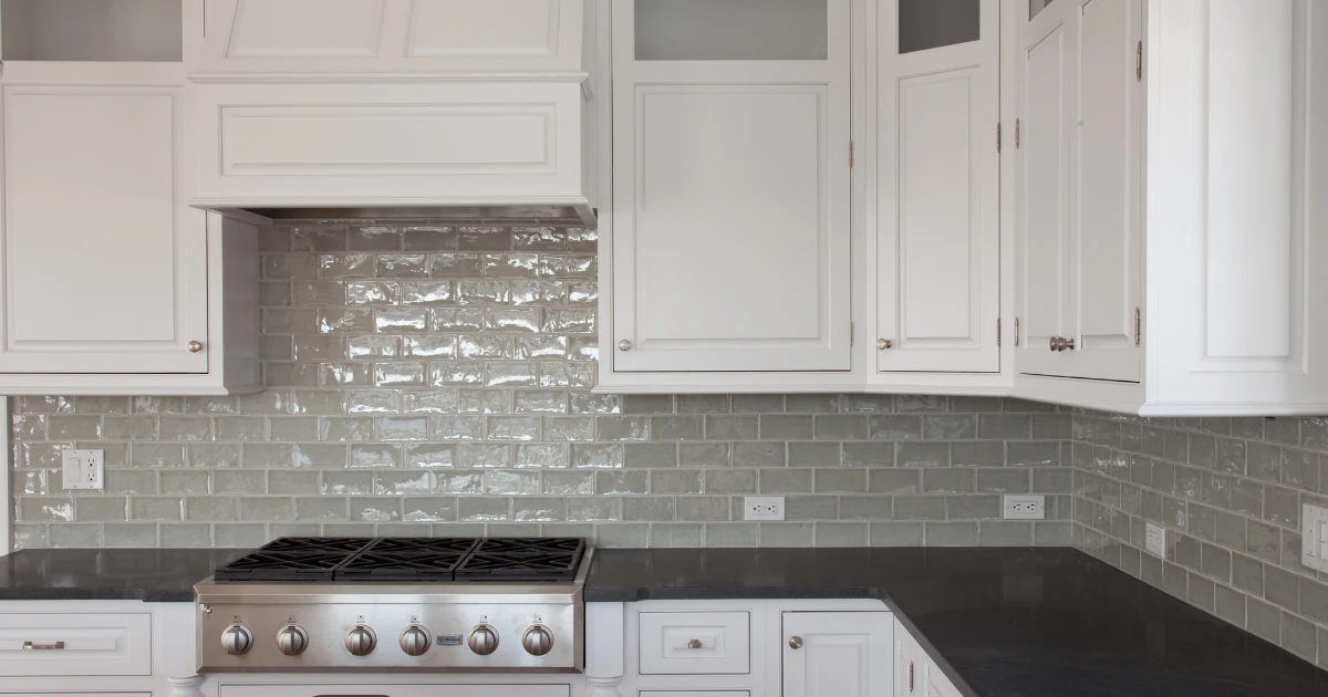 A finished white kitchen with painted cabinetry, a gray subway tile backsplash, a stainless steel gas range hood, and black countertops.
