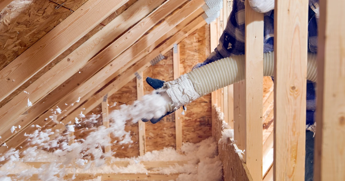 A worker in an attic blows loose-fill insulation into wall framing between roof rafters using a large hose.