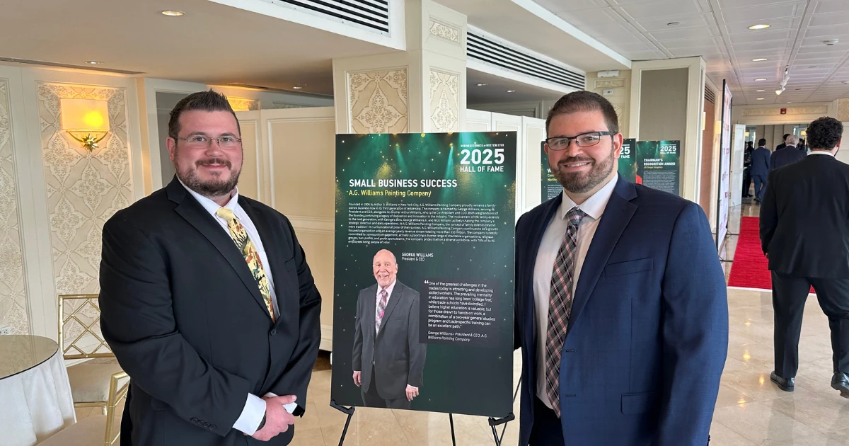 Two men in suits stand beside an easel-mounted sign reading "Small Business Success, A.G. Williams Painting Company" with a photo of an older man in a suit and a "2025 Hall of Fame" logo.