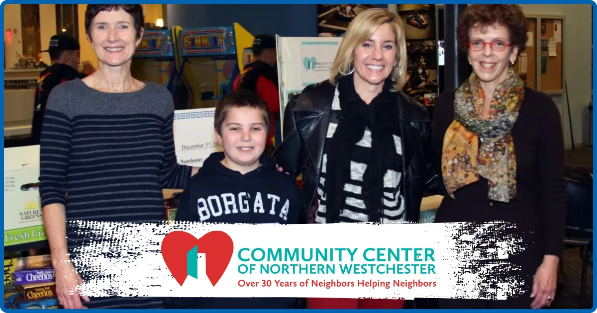 Two women, a boy, and another woman pose together indoors in front of a banner reading 'Community Center of Northern Westchester, Over 30 Years of Neighbors Helping Neighbors.'