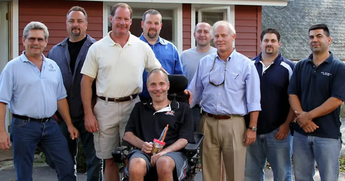 Group photo of nine men, including one seated in a wheelchair in front, standing outside a red house with white trim.