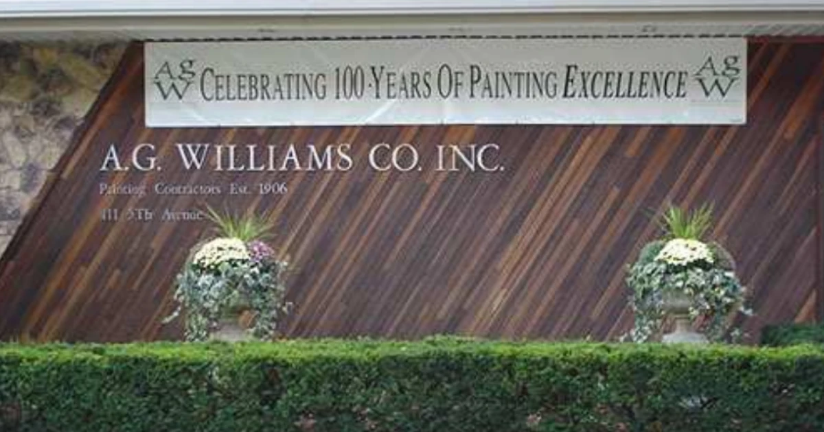 Exterior sign for A.G. Williams Co. Inc. on a wood-paneled building, with a banner above reading "Celebrating 100 Years of Painting Excellence" behind trimmed hedges and potted flowers.