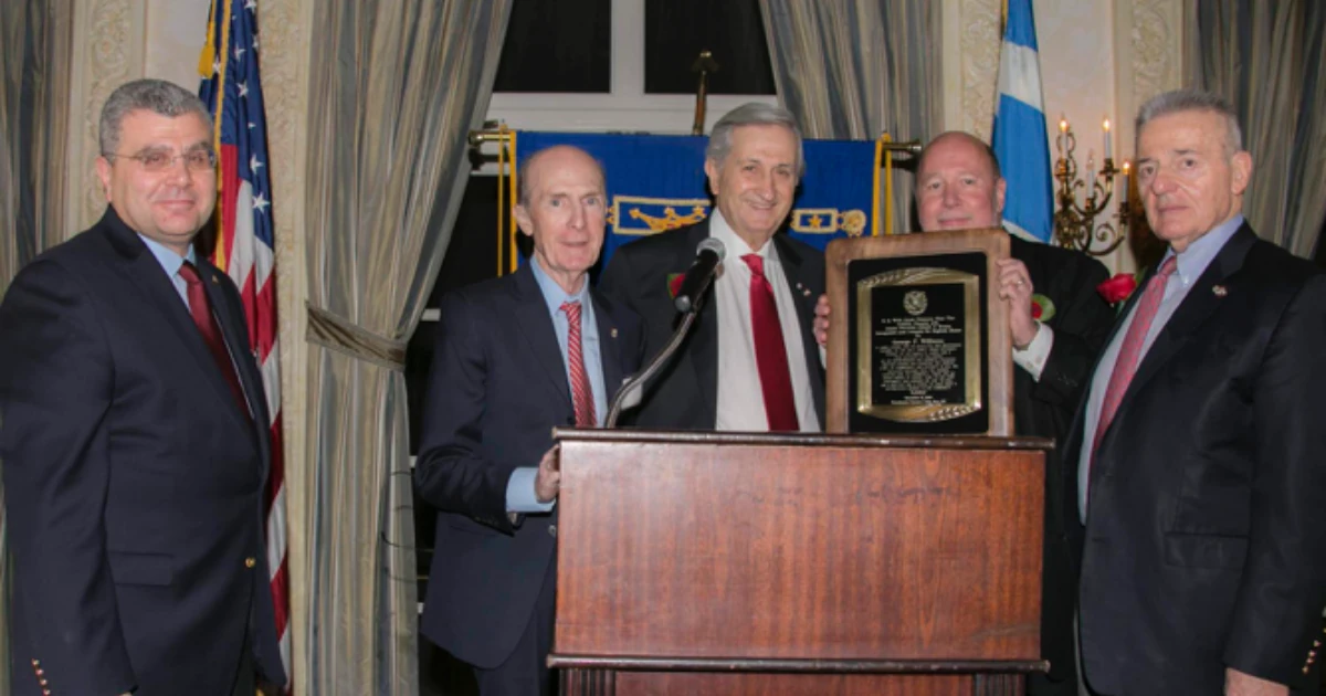 Five men in suits standing at a podium, one holding a plaque award, in front of American and blue-and-white flags at an indoor event.