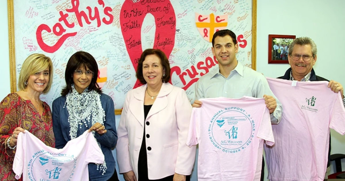 Five people stand in front of a signed pink ribbon banner reading "Cathy's Crusaders," with four of them holding folded pink A.G. Williams Painting Company T-shirts for a Support-A-Walk breast cancer fundraiser.