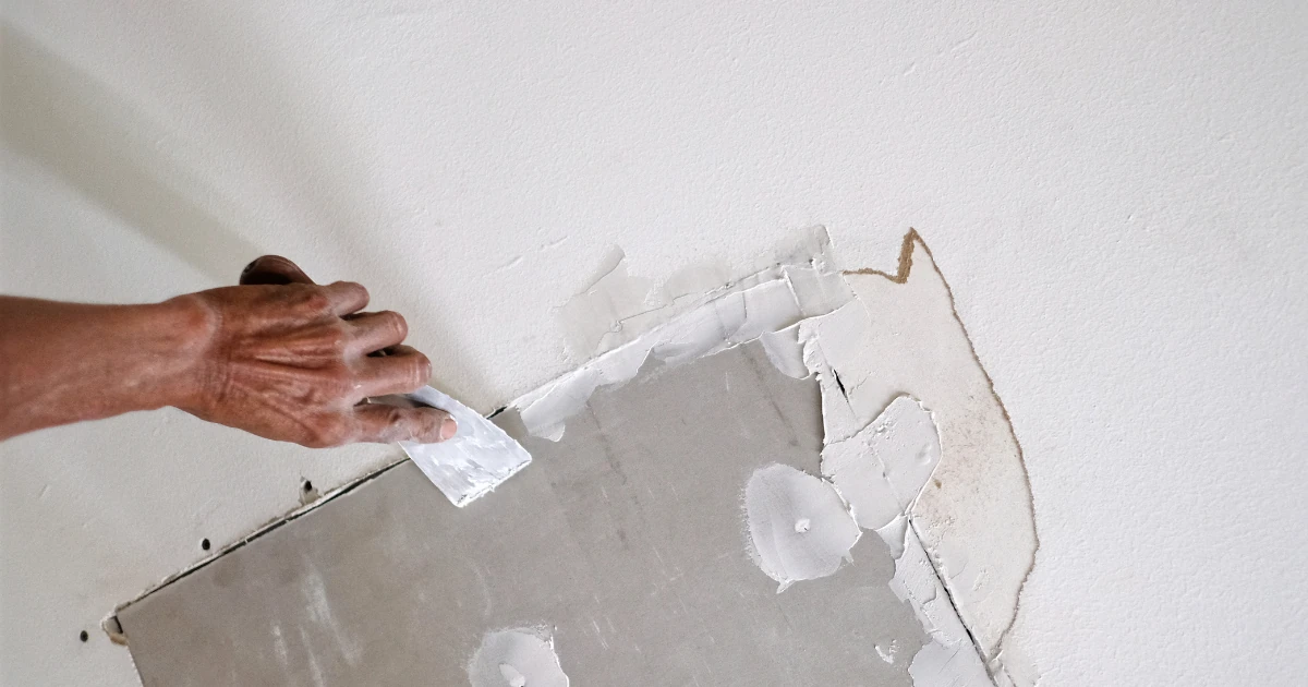A hand uses a putty knife to spread joint compound over an exposed drywall patch on a white wall.