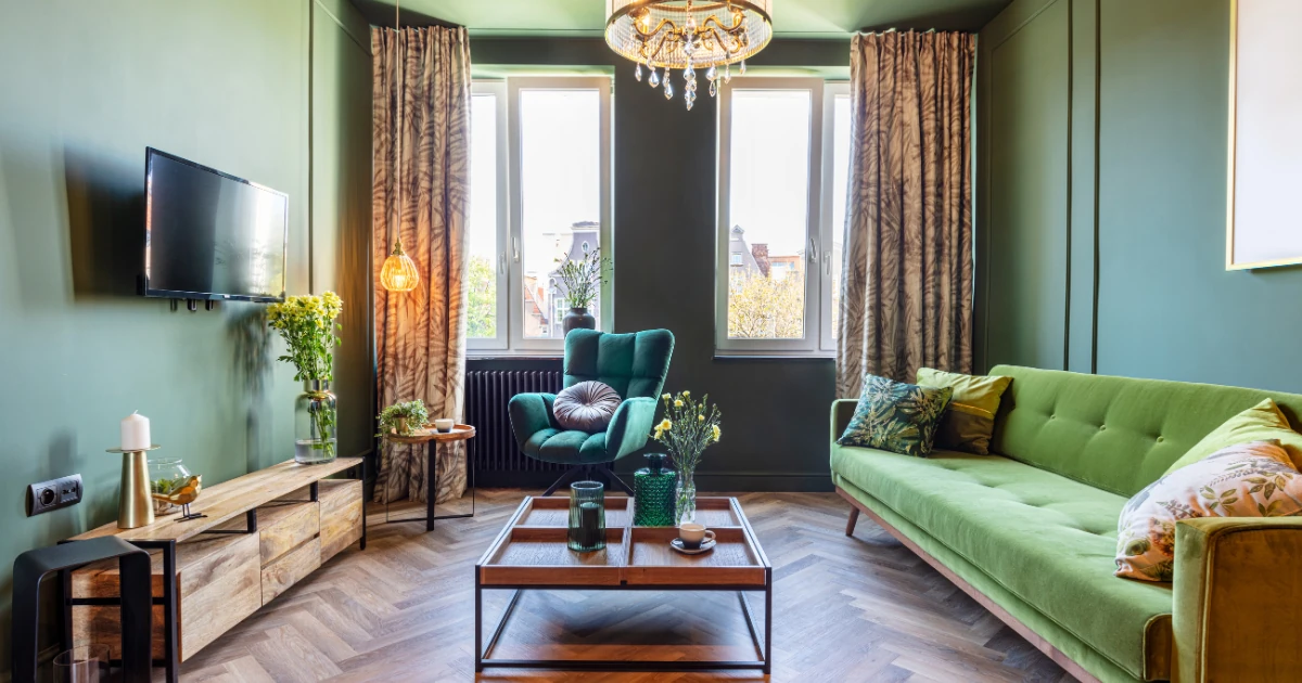 A living room fully painted in matching sage-green walls, featuring a green tufted sofa, a teal accent chair, patterned curtains, a herringbone wood floor, and a crystal chandelier.