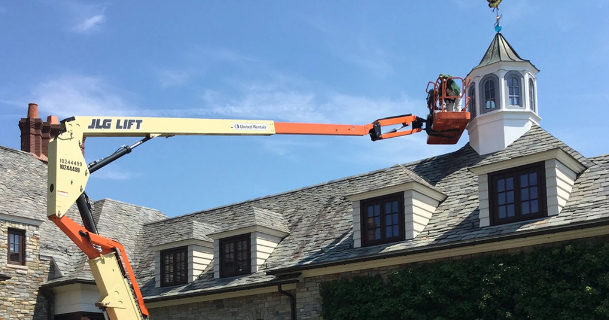 A worker in an orange boom-lift basket reaches a white cupola atop a slate-roofed stone house under a clear blue sky.