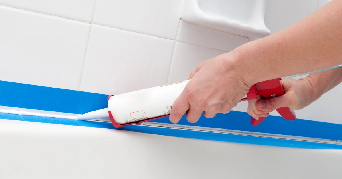 A hand using a caulking gun to apply a bead of caulk along a bathtub edge next to blue painter's tape and white tile.