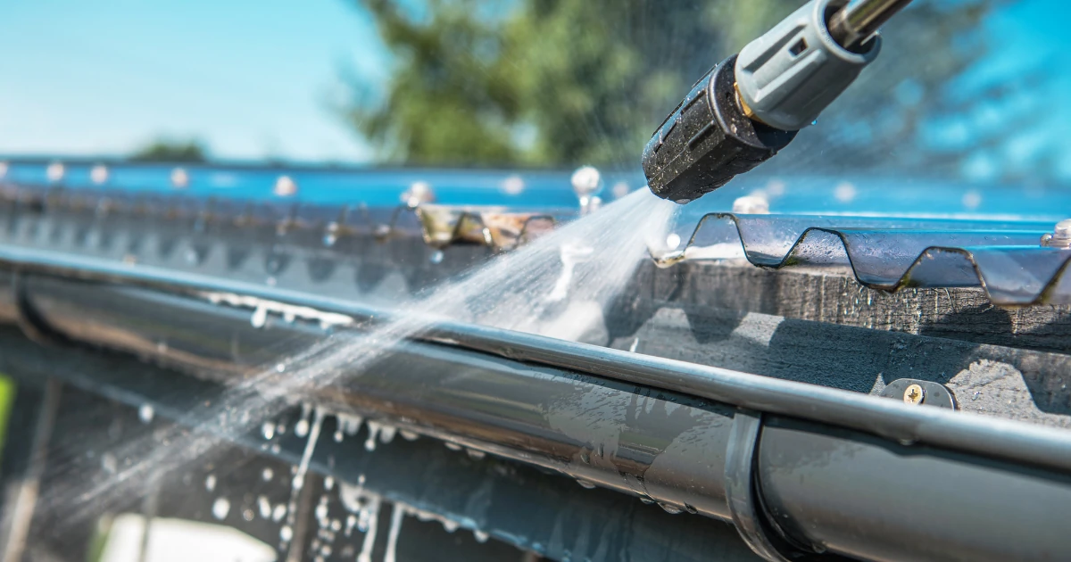 A pressure washer nozzle sprays water onto a gutter and clear plastic roof panel to clean it.