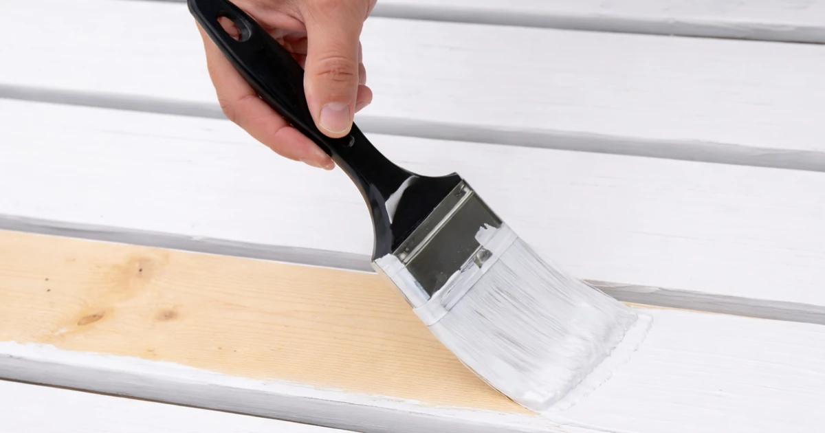 Close-up of a hand using a paintbrush to apply white paint to a bare wood board.