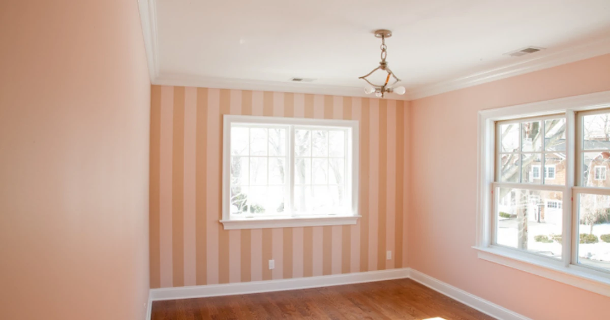 An empty room with pink walls, a striped pink-and-cream accent wall, crown molding, and two windows.