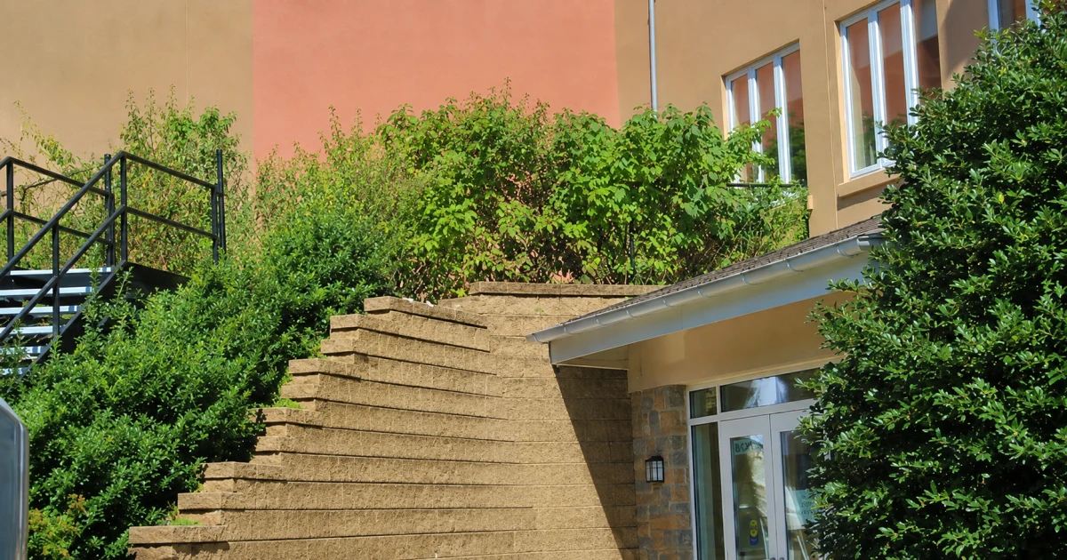 Tan stucco condominium exterior with landscaped retaining walls, an exterior stair, and mature greenery.