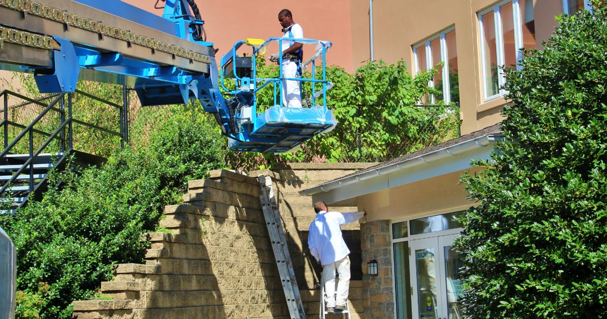 Two painters, one on an aerial boom lift and one on a ladder, painting the exterior of a stucco commercial building surrounded by greenery.