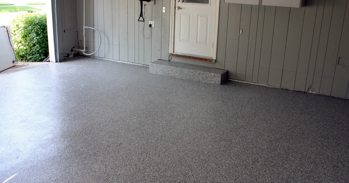 A garage interior with a dark gray speckled epoxy floor, a white entry door with a small step, and wood-paneled walls, with the overhead door open to a yard.