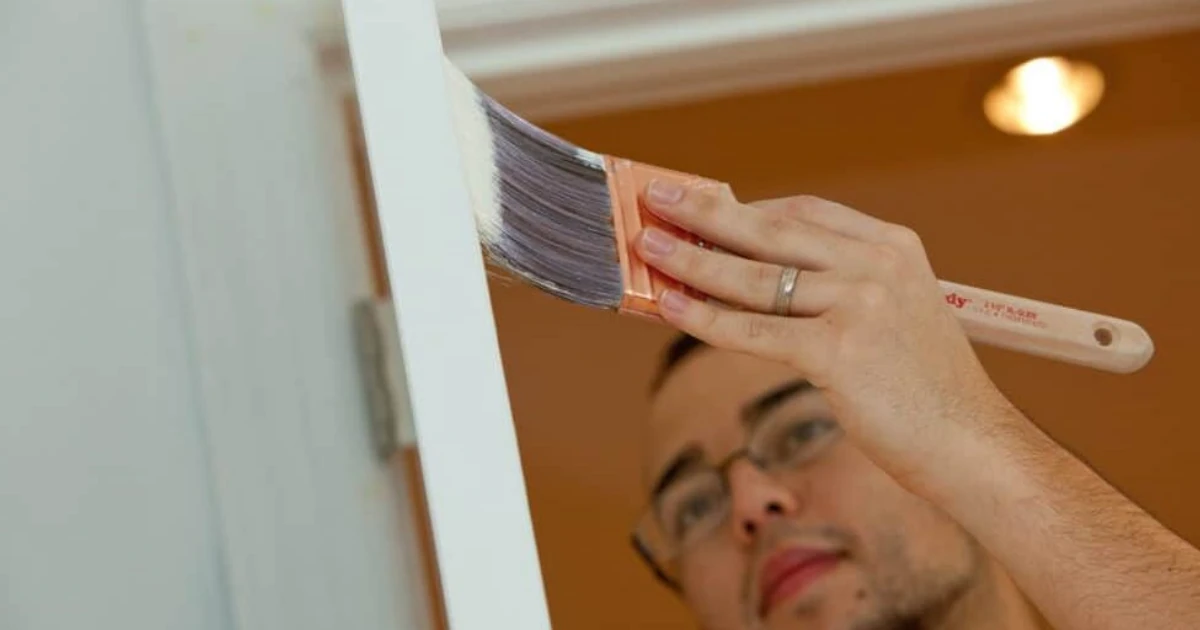 Close-up of a hand holding a paintbrush applying paint to a white door frame.