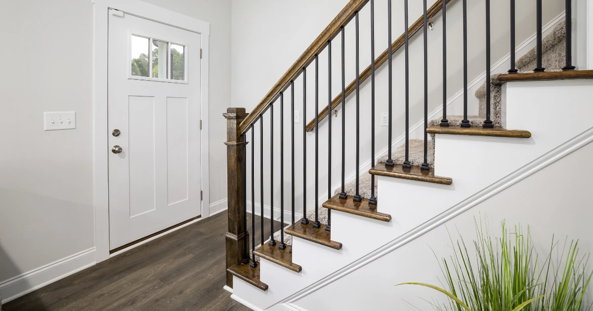 A bright entryway with a white front door, dark wood stair treads, black metal balusters, and a wood handrail.