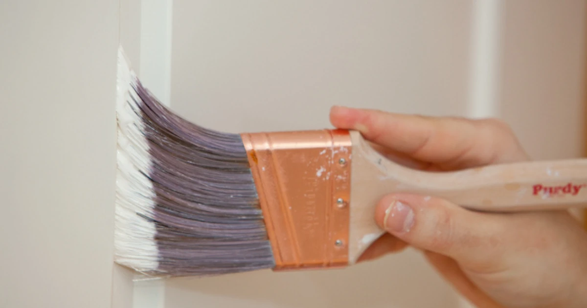 A close-up shows a hand holding a Purdy paintbrush loaded with white paint applying it to a wooden door frame.