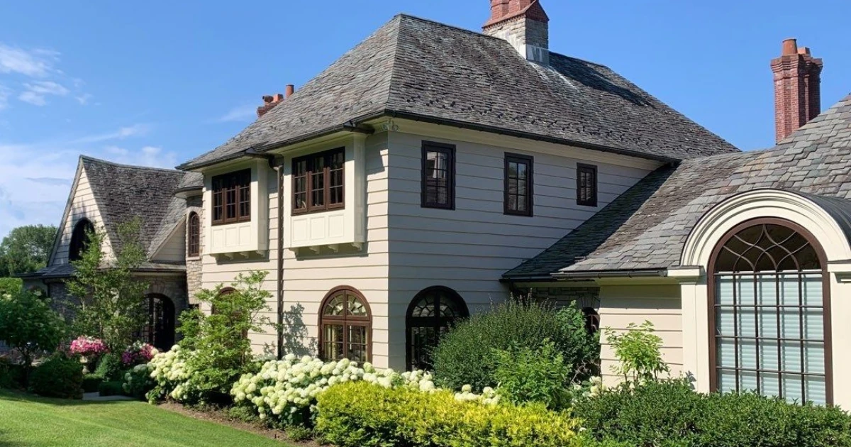 The exterior of a large house with cream-colored siding, dark-trimmed windows, a slate-look shingle roof, brick chimneys, and landscaped shrubs and hydrangeas.