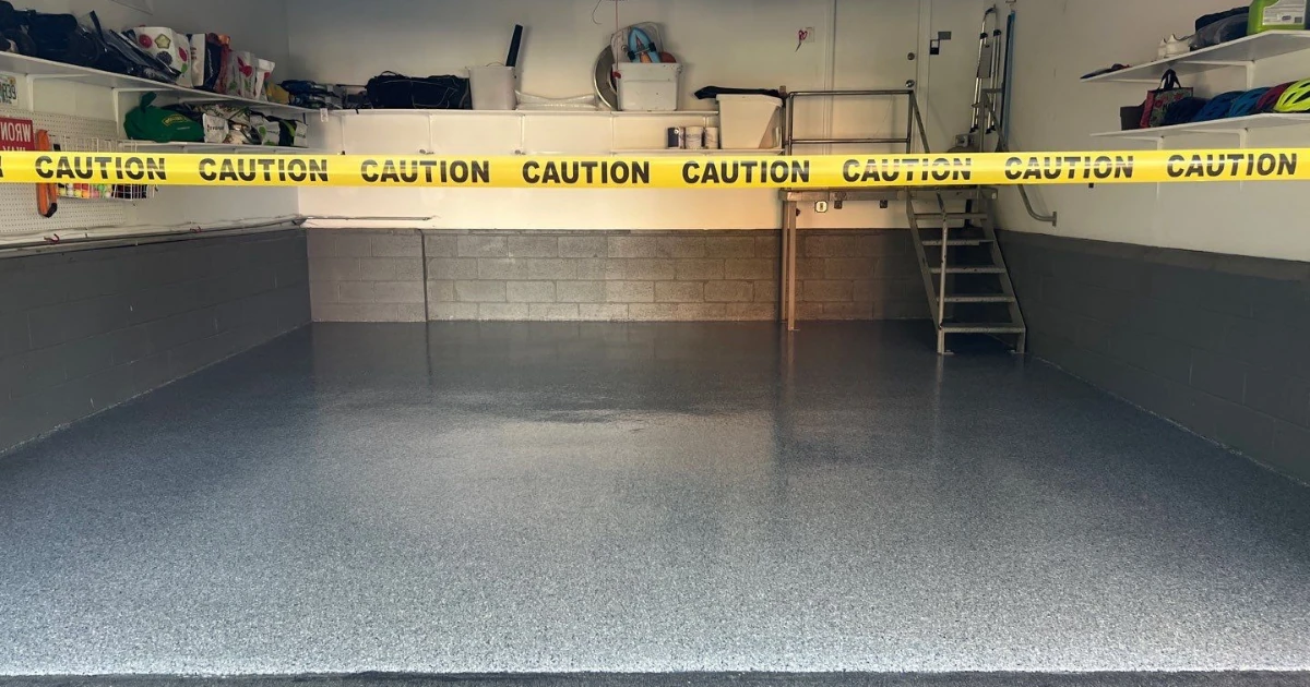 An empty garage with a gray speckled epoxy floor cordoned off with yellow caution tape, showing wall shelving, a small platform, and steps along the back wall.