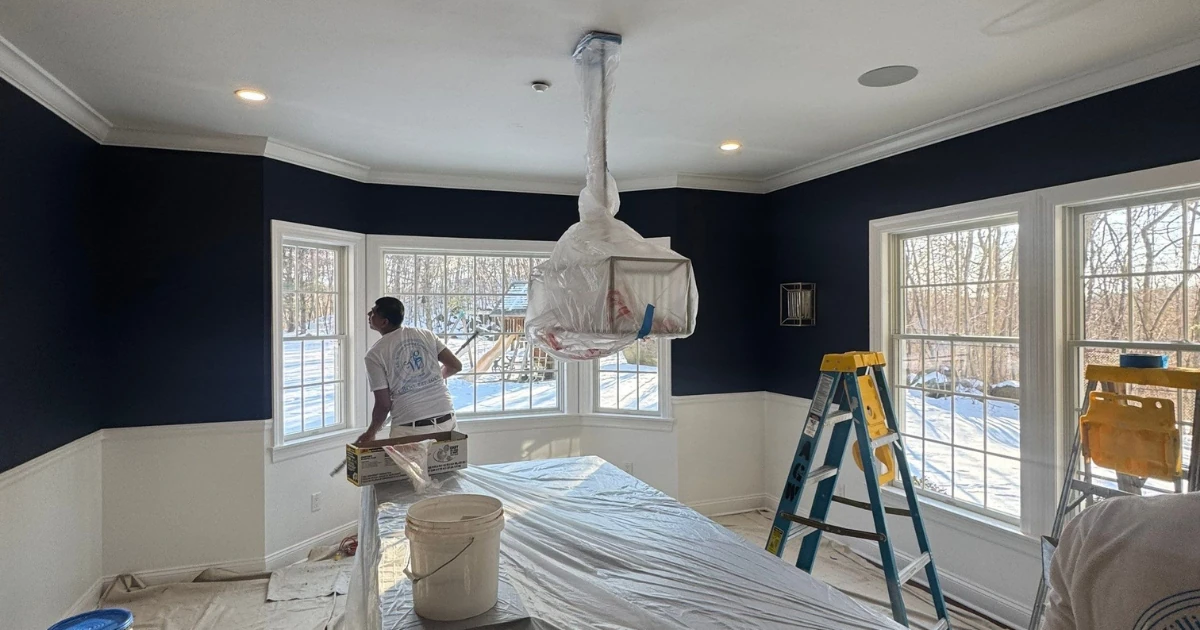A room mid-painting with dark navy walls and white wainscoting, a chandelier wrapped in plastic hanging from the ceiling, drop cloths covering furniture, paint buckets, and step ladders near bay windows overlooking a snowy yard.
