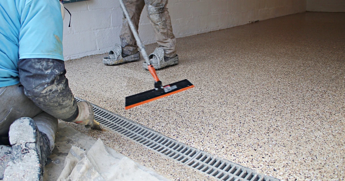 Two workers in a garage apply a speckled epoxy flake floor coating, one spreading it with a long-handled tool and the other detailing the edge near a floor drain.