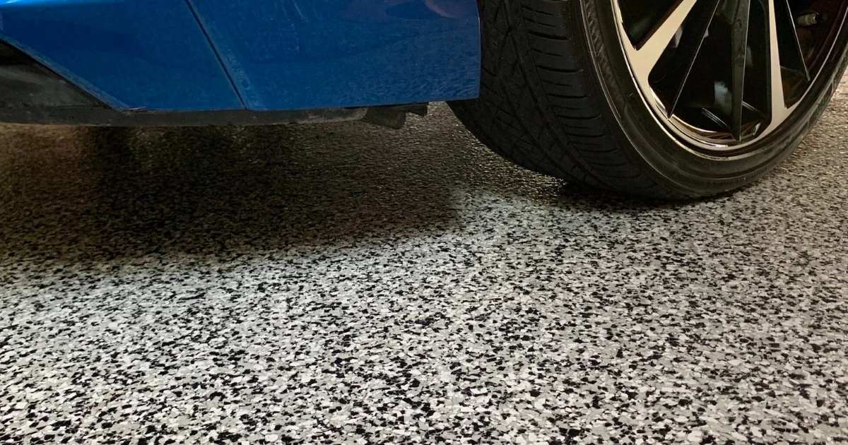 A close-up of a blue car's rear wheel and bumper resting on a gray-and-white speckled epoxy garage floor coating.