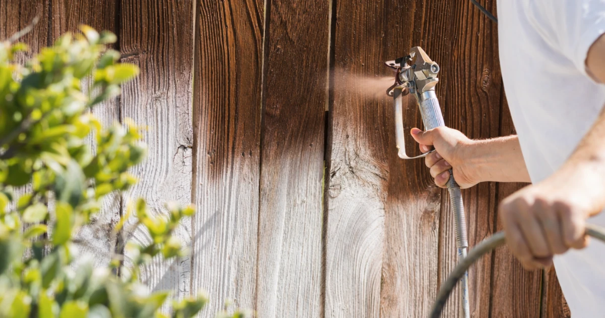 A person's arm and hand spraying wood stain or paint onto a weathered wooden fence using a handheld sprayer.