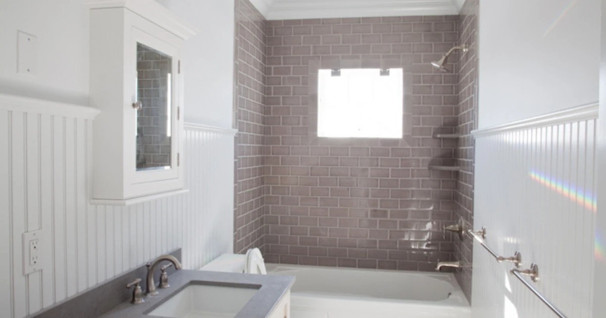 A bathroom with white beadboard wainscoting, a mauve subway-tile shower surround with a window, a white tub, and a gray-topped vanity.