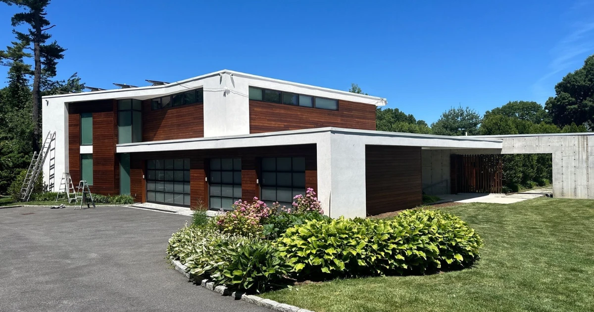 A modern two-story house with white stucco and dark wood paneling, flat roofline, and landscaped garden, with ladders leaning against the facade.