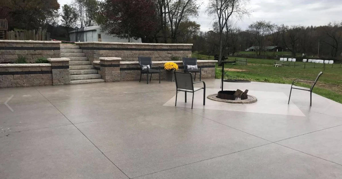 Outdoor stamped concrete patio with a stone retaining wall and steps, patio chairs, potted yellow mums, and a circular in-ground fire pit, with a rural landscape in the background.