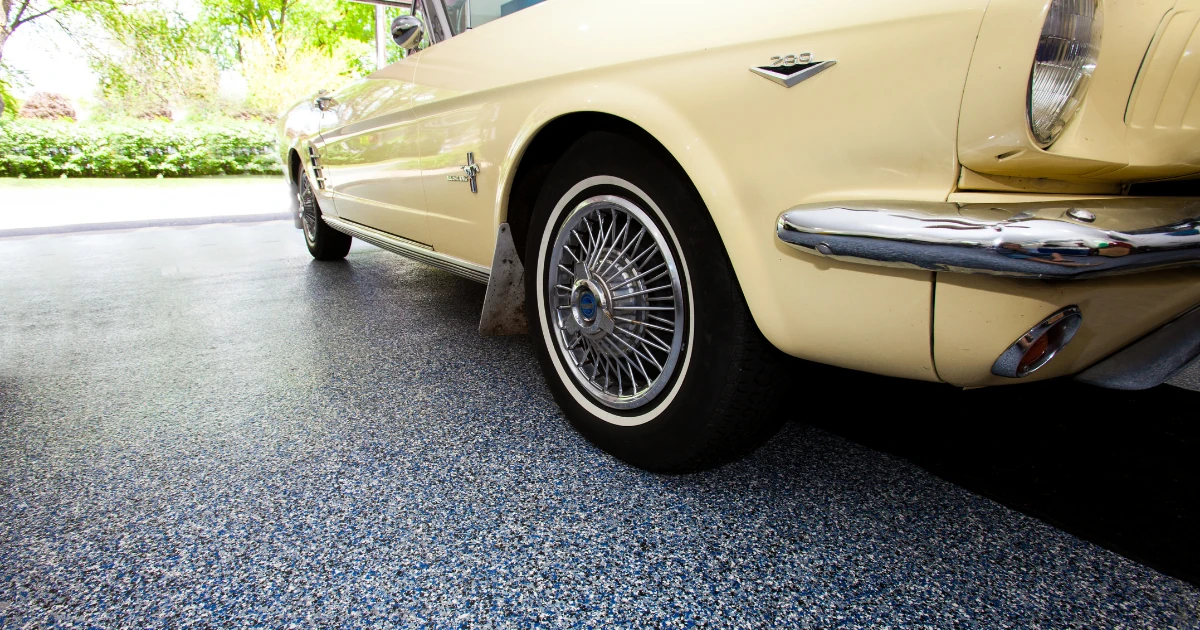 A close-up of a cream-yellow vintage car with wire-spoke wheels parked on a gray speckled epoxy garage floor coating.