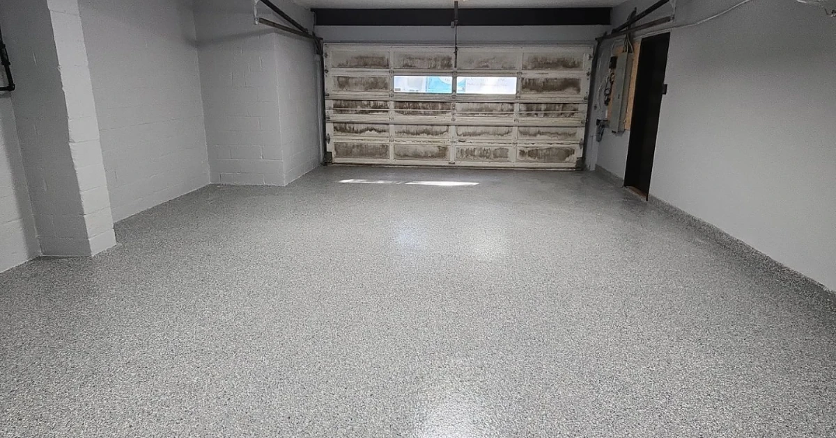 An empty two-car garage with a newly finished gray speckled epoxy floor and a weathered white garage door at the far end.