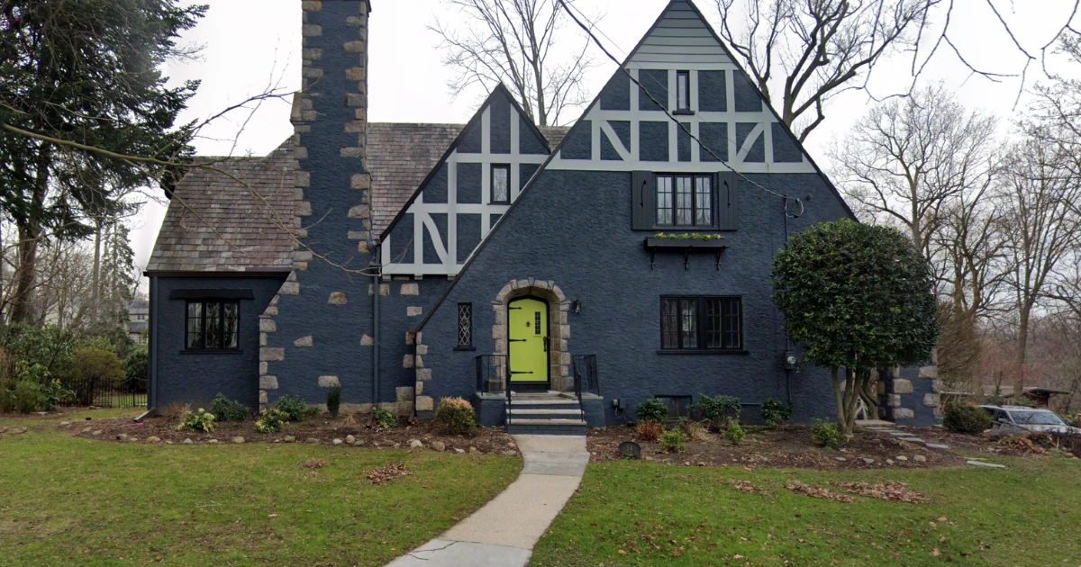 A Tudor-style stone-and-stucco house painted dark blue with light gray half-timbering and a bright chartreuse front door, seen from the front lawn.