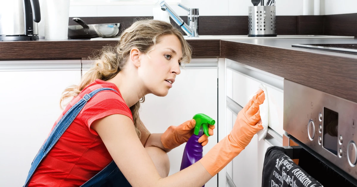 A woman wearing orange rubber gloves wipes a white kitchen cabinet drawer with a cloth while holding a spray bottle.