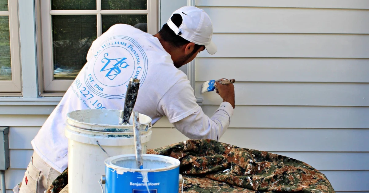 A.G. Williams painter brushing gray exterior siding beside a window, with protected paint supplies in the foreground.