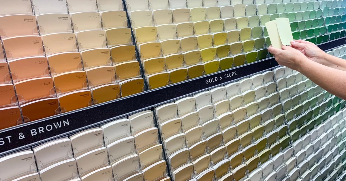 A hand holding two paint chips against a large in store display of neutral, gold, and green paint color cards.