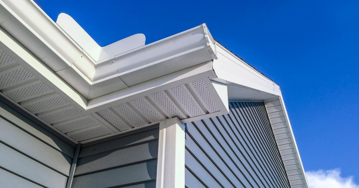 Upward view of a roofline showing crisp white painted fascia, vented soffit, and gutter above gray siding against a clear blue sky.