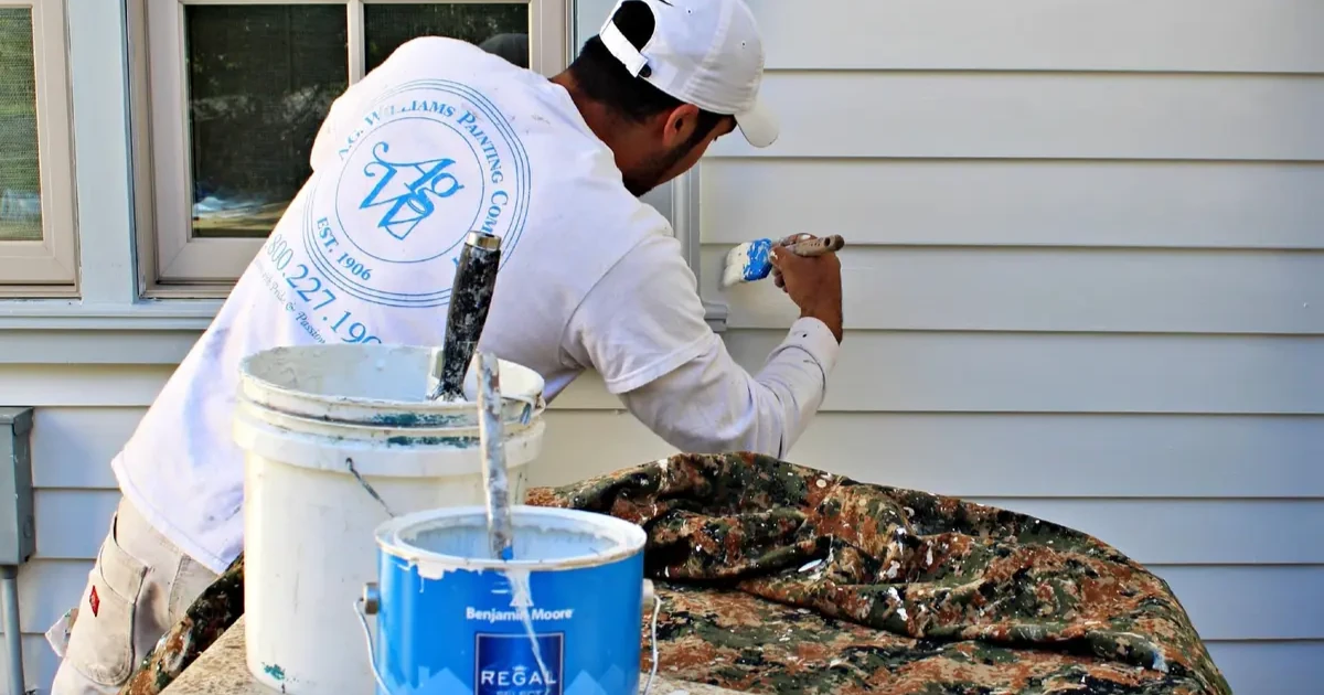 An A.G. Williams painter in a branded shirt brushing paint onto lap siding beside buckets of premium exterior paint.