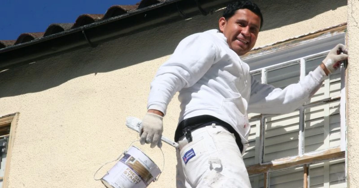 A painter on a ladder cutting in a window frame by brush, holding a bucket of exterior primer against a stucco wall.