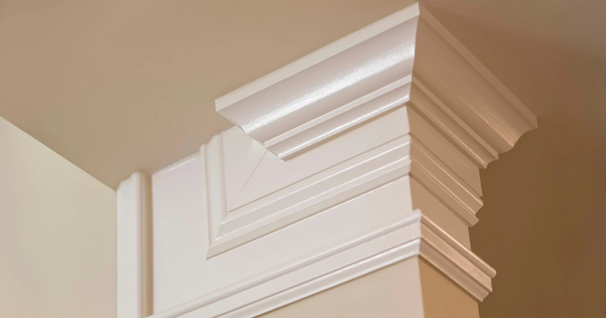 Close-up of layered white crown moulding installed where a beige wall meets the ceiling, showing stacked profiles at an outside corner.