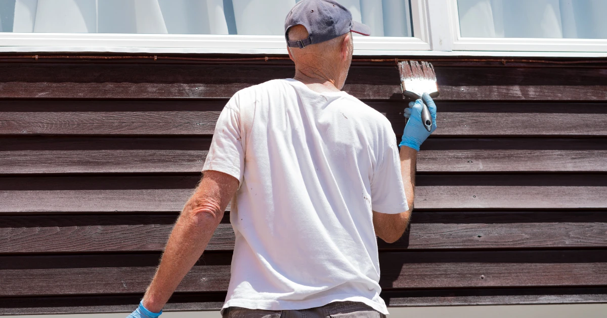 A painter brushing a coat of finish onto horizontal wood siding boards beneath a white window frame.