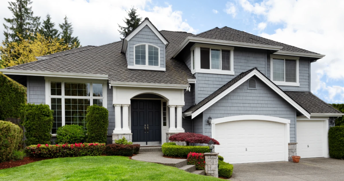 A gray shingled two story home with white trim, a dark front door, and flowering beds under a bright blue sky.