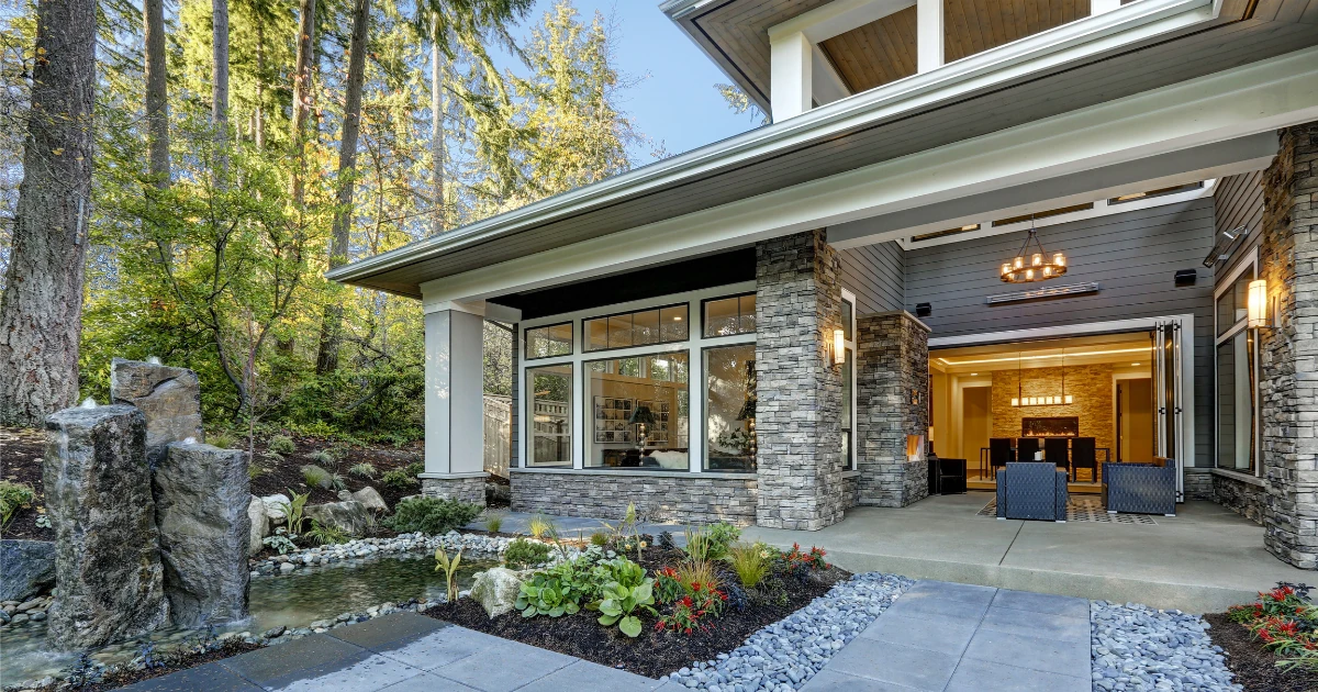 A modern home exterior with gray siding, stone columns, and warm interior lighting visible through large windows at dusk.