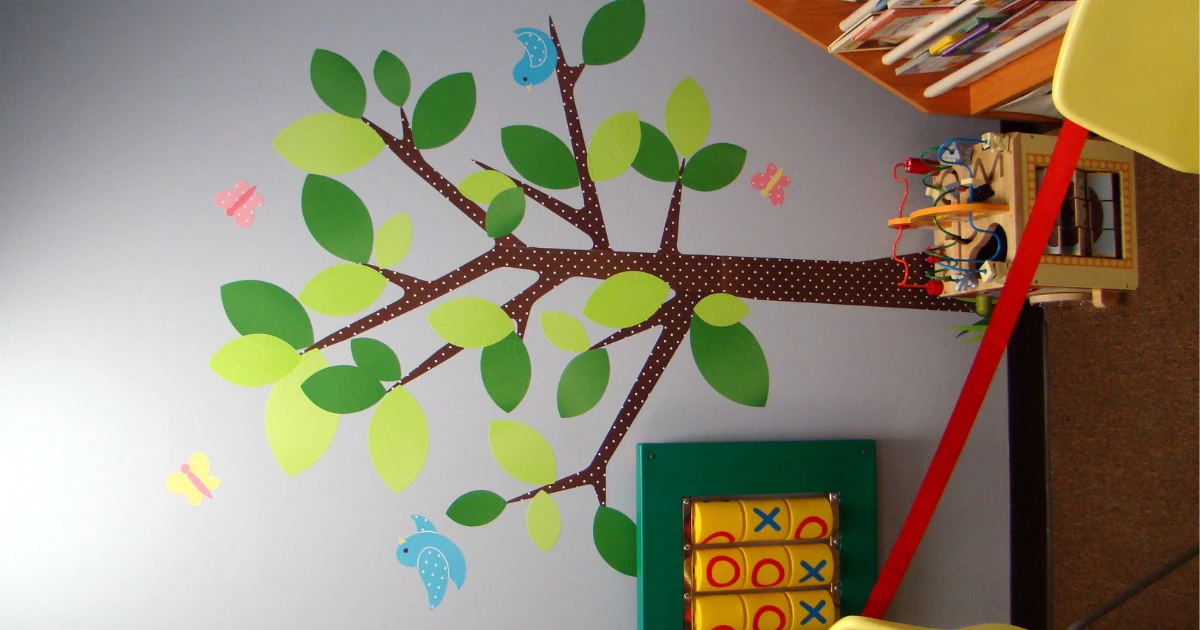 Hand painted tree mural with green leaves, blue birds, and butterflies on a gray wall beside a children's play area in a nonprofit reception room.
