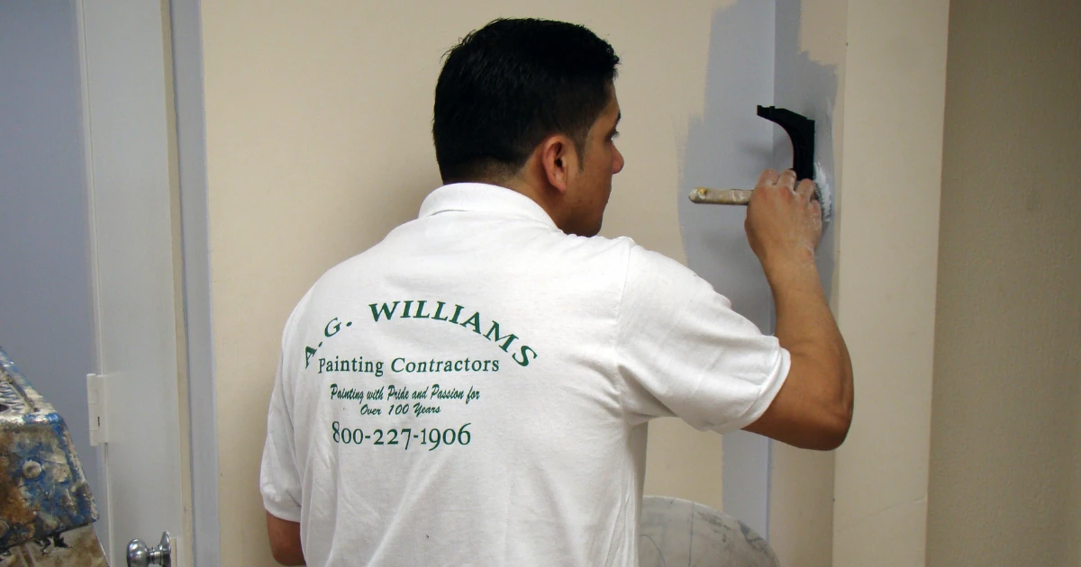 An A.G. Williams painter in a branded company shirt rolling fresh paint onto a reception area wall.