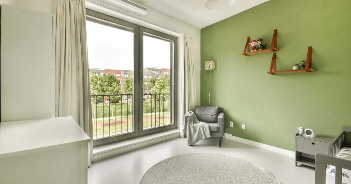 Nursery with a green painted accent wall, shelves, crib, chair, and large window.