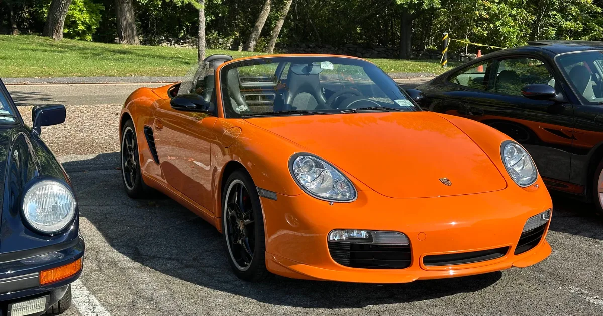Orange Porsche Boxster convertible parked between classic cars at Cars & Coffee.
