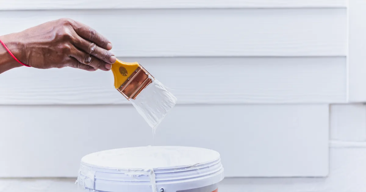 Painter holding a brush coated in white primer above an open bucket.