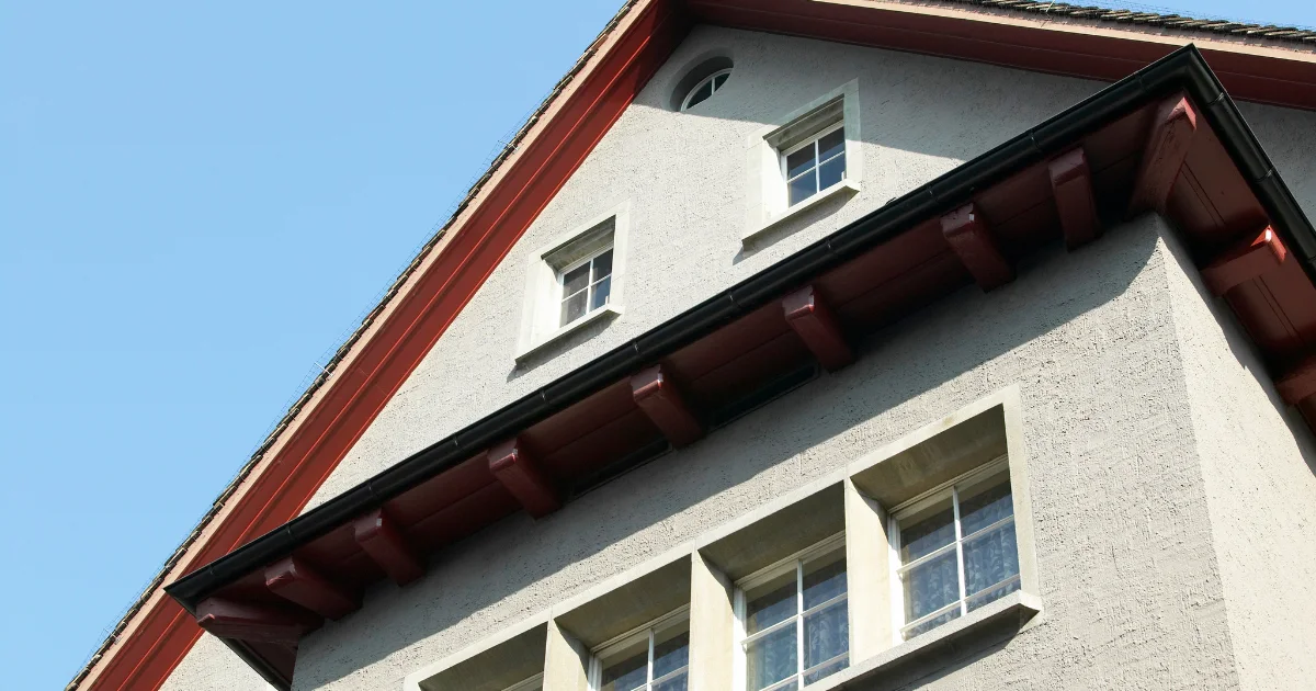 White stucco house exterior with red roof trim and black gutters viewed from below.