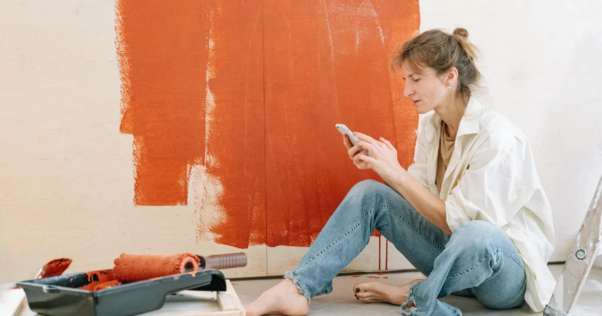 Person seated beside painting supplies and a ladder in front of a partly painted orange wall.